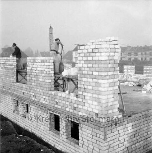 Wohnungsbau: Neubaugebiet Kellerberg-Süd: Wohnblock im Bau mit Grundmauern: darauf Arbeiter: links Lastenaufzug: hinten rechts und Mitte Blocks vor Fertigstellung und im Rohbau mit Gerüst: hinten links Einfamilienhäuser