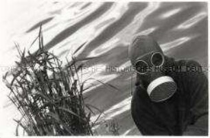 Mann mit Gasmaske sitzt vor einem Gewässer, rechts von ihm Schilfbüschel (Altersgruppe 14-17)