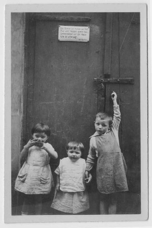 Kleinkinder vor einem Tor, daran ein Schild mit der Aufschrift "Das Spielen der Kinder auf Hof, FLur und Treppen sowie das umherstehen vor der Haustüre sind untersagt! Der Besitzer