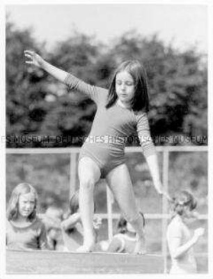 Mädchen in Gymnastikanzug balanciert auf einem Balken, im Hintergrund weitere Mädchen und Bäume (Altersgruppe bis 14)