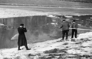 Zugefrorener Elsterflusslauf : Zwei Männer mit einer Stange beim Abfischen von durch Eissprengungen getöteten Fischen am zugefrorenen Elsterflusslauf. Ein Mann in Uniform fotografiert die Szene.