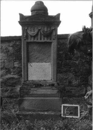 Grabstein 6 (Index: 100080725) Material: Sandstein, weiße Marmorplatte Zustand: Platte gesprungen, Aufsatz beschädigt Maße HxBxT: 165x49x27 cm Beschreibung: Stele, getreppter Sockel, Giebel mit Aufsatz, rechteckige Schriftplatte: Grabstein 6