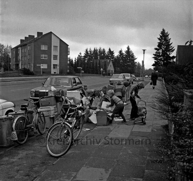 Gerhart-Hauptmann-Straße: Sperrmüll: Kinder beim Durchwühlen der Haufen: hinten links Wohnblock, Straßeneinmündung: rechts Zaun, Vorgarten: Fahrradweg, Straßenlampen, Fahrräder, Mülltonnen, parkende Autos, hinten Fußgänger