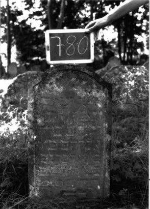 Grabstein 780 (Index: 100104691) Material: Sandstein Zustand: stark verwittert Maße HxBxT: 84x45x10 cm Beschreibung: Stele, Konvexbogenabschluss, Schriftfeld eingetieft, im Bogenfeld segnende Hände Symbolik: segnende Hände: Grabstein 780