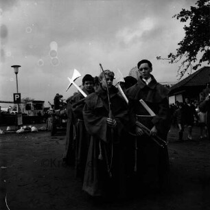 Karpfenfest: Umzug: Fußgruppe, Mönche in Kutten, mit Zirkel, Bogen, Äxten, Kescher: im Hintergrund Traktor, Straßenlampe, Schild Parkplatz, Zuschauer, 8. Oktober 1967