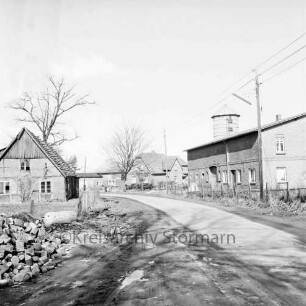 Dorfstraße mit Bauernhäusern und Silo: links Haufen mit Holzscheiten