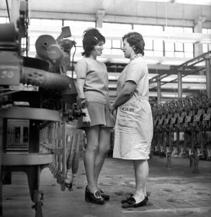Frauen in der Spinnerei : Zwei Frauen, eine junge in Freizeitmode und eine alte in Arbeitskleidung, unterhalten sich in einer Spinnmaschinenhalle