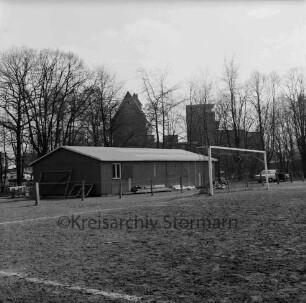 Trave-Stadion: Exer: vorn Fußballplatz mit Tor: dahinter Zaun, Holzbaracke, Bretterstapel und Pkw: dahinter Bäume und Silo der Johannes Ströh Mühlenwerke, "Gloria-Mühle"