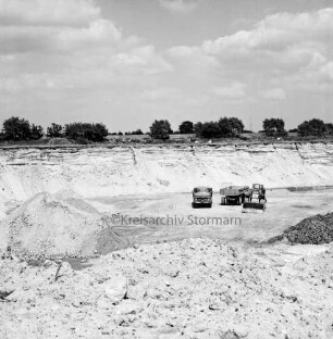 Kiesgrube: hinten rechts Lkw, Bagger und Anhänger auf Grubengrund: vorn Sand- und Kieshaufen: im Hintergrund Bäume und Felder