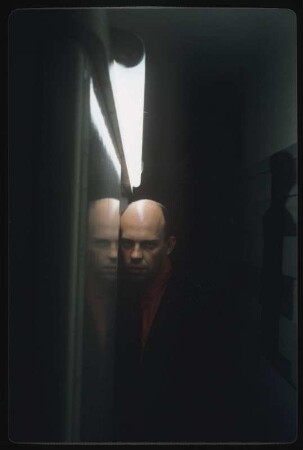 Selbstportrait Ingolf Thiel : Ingolf Thiel im schwarzen Jacket und roten Hemd in einem engen Flur, beleuchtet durch eine Neonröhre, mit dem Kopf an einen Rahmen gelehnt; auf der anderen Wand hängt eines der Fotos aus Thiels Serie "Bandagierte Körper"