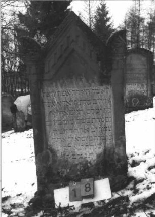 Grabstein 18 (Index: 99608702) Material: Sandstein Zustand: leicht verwittert Maße HxBxT: 115x55x12 cm Beschreibung: Stele auf Sockel, eingezogener Spitzgiebel mit Eckakroterien, vertiefter Textteil in gleicher Form mit Stalagtitengebälk: Grabstein 18
