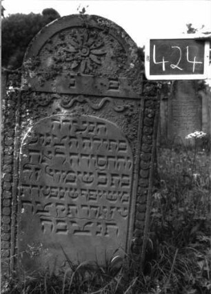 Grabstein 424 (Index: 100102774) Material: Sandstein Zustand: gut erhalten Maße HxBxT: 129x62x12 cm Beschreibung: Stele, eingezogener Rundbogenabschluss, im Bogen Blüte, Schriftfeld erhaben mit Korbbogenabschluss, darüber Wellenlinie, auf seitlichen Rahmen Scheibenfries Symbolik: Blüte, Wellenlinie, Scheibenfries: Grabstein 424