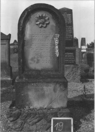 Grabstein 19 (Index: 100113959) Material: Sandstein Zustand: leicht verwittert Maße HxBxT: 150x60x20 cm Beschreibung: Stele, getreppter Sockel, unterer Sockel rusticaartig, Rundbogenabschluß Symbolik: Blüte: Grabstein 19