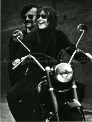 Lachendes Paar (Jürgen Klauke und Hannelore Wiese) mit Sonnenbrillen sitzt auf einem Motorrad