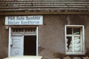Lassan. Haustür mit Inschrift "PGH 'Gute Qualität' Bäcker Konditoren" und Fenster mit dahinter gestapelten Mehlsäcken