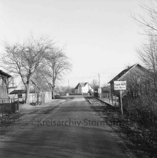 Ortseingang mit Dorfstraße: rechts Ortsschild: im Hintergrund Bauernhäuser und Bäume