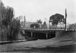 Brücke : Eine Brücke, über die eine Straßenbahn fährt, über einen Kanal an einer Parkanlage. Im Hintergrund hohe Gebäude.