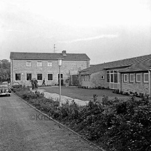 Ahrensburg: Reeshoop: Einweihung: Außenaufnahme: links Wirtschaftsgebäude, rechts Wohnflügel: Ankunft der Gäste, Autos: davor Grünanlage, mit Rasen, Sträuchern, Plattenweg: hinten Kunst am Bau, Bronzeplastik "Ziehharmonikaspieler" von Bildhauer Karlheinz Goedtke: vorne links Zufahrt, 25. Oktober 1961