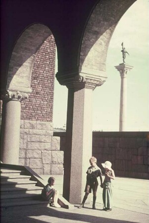 Drei Kinder an einer Treppe unter einer Arkade. Blick zu einer Säule mit Fahnenträger?