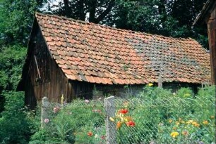 blühende Pflanzen in einem Nutzgarten, dahinter Wirtschaftsgebäude