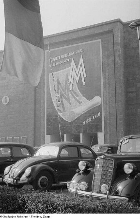 Parkende PKW auf dem Gelände der Leipziger Messe 1954, im Hintergrund Werbung für die Frühjahrsmesse 1955
