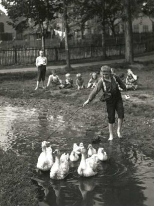 Kind als Gänsehirt im Dorfbach und ihm zuschauende Kindergruppe auf der Wiese