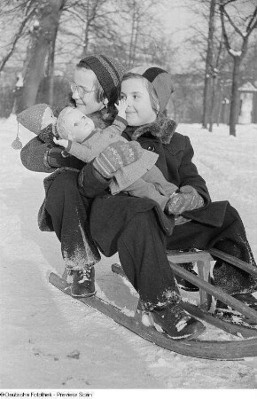 Zwei Kinder mit Puppen auf dem Arm auf einem Schlitten in einer Schneelandschaft