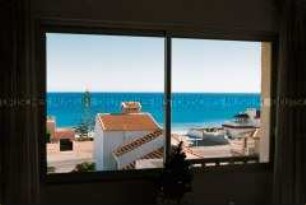 Blick aus einem Fenster auf ein Haus und das dahinterliegende Meer (Altersgruppe bis 14)