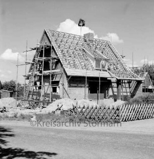 Lasbek-Dorf: Bau eines Einfamilienhauses: Rohbau mit Richtkrone und Gerüst: davor Baumaterial: vorn rechts Jägerzaun: im Vordergrund Straße: hinten rechts weiterer Rohbau