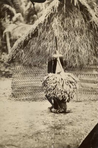 "Duk Duk representative in front of a mat-roofed hut, New Britain"