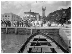 Barbados. Hafenszene mit Einheimischen und Touristen auf der Kaimauer, im Hintergrund eine Kirche. Blick aus einem vertäuten Ruderboot