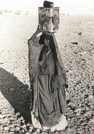 Ghat, Libyen. Tuaregfrau (Targia) in volkstypischer Kleidung mit Benzinkanister als Wasserbehälter, auf dem Kopf tragend