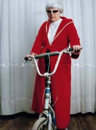 Ältere Frau mit Sonnenbrille und rotem Bademantel auf einem kleinen Fahrrad (Altersgruppe 18-21)