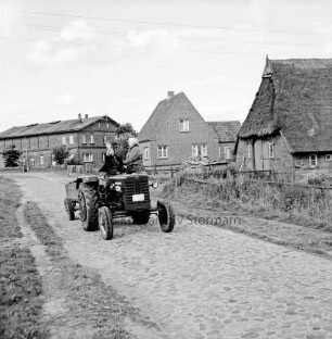 Dorfstraße mit Kopfsteinpflaster: im Vordergrund landwirtschaftliches Fuhrwerk mit winkender Fahrerin: im Hintergrund Wohnhaus und Bauernhäuser