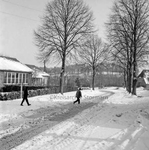 Dorfstraße mit zwei Kindern: links Volksschule mit Vorgarten: hinten Bäume und Häuser: Winter