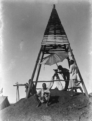 Vermessung : Ein Vermesser bei der Arbeit an einem Messinstrument unter einem pyramidenförmigen Holzbau. Ein Sonnenschirm spendet ihm Schatten. Unter dem Messinstrument ein Lot. Um ihn herum drei weitere Personen. Links im Hintergrund ein Zelt mit einer Flagge.