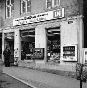 Willfang Buchhandlung: Hude 7: gleichzeitig Geschäftsstelle der Lübecker Nachrichten LN: Außenaufnahme: Eingang mit Zeitungsständer, Werbeaufsteller: Schaufenster, darüber Firmenschild: an Außenwand Zigarettenautomat, Auslage-Kasten, Straßenschild: Fußgänger: rechter Bildrand Parkuhr