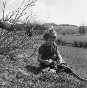 Kinderbilder. Kleines Mädchen, auf einer Wiese beim Blütenkranzflechten sitzend