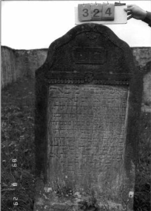 Grabstein 324 (Index: 100107748) Material: heller Sandstein Zustand: stark verwittert, teilweiser Textverlust Maße HxBxT: 176x41x14 cm Beschreibung: Stele, Abschluß Sonderform (geschwungen eingezogener flacher Kleeblattbogen), Schriftfeld rechteckig eingetieft Symbolik: Blüte, Halbblüte, Tropfenband: Grabstein 324
