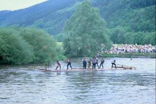 großes Floß mit Flößern beim Staken und Steuern (Steuermann am Ende), Publikum am Ufer