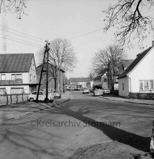 Hauptstraße: rechts und links hinten Wohn- und Geschäftshäuser: davor parkende Pkws und Lieferwagen: links Strommast