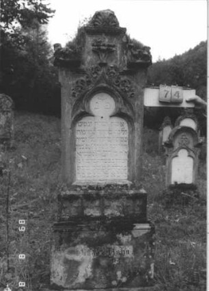 Grabstein 74 (Index: 99799555) Material: Sandstein, Schriftplatte Marmor Zustand: verwittert Maße HxBxT: 197x61x26 cm Beschreibung: Stele, Mittelerhöhung, Verdachung, Mittelpalmette, eingelassene Schriftplatte mit Kleeblattbogenabschluß, getreppter Sockel Symbolik: floral: Grabstein 74