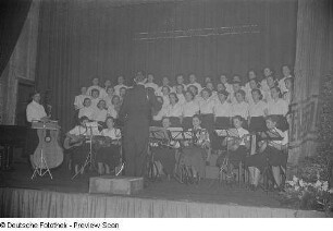 Chor, Chorleiter und Instrumentenensemble während eines Auftritts beim Bezirksausscheid der Volkskunstgruppen 1954