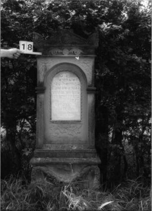 Grabstein 18 (Index: 100100624) Material: Sandstein mit weißer Marmorplatte Zustand: Sockel verwittert Maße HxBxT: 167x60x28 cm Beschreibung: ädikulaartige Stele mit Pilastern, Sockel, Giebel und Akrotere, eingetiefte Schrifttafel mit Rundbogen Symbolik: Blüten, Blätter, Zweige, um die Schrifttafel laufendes Fries, Girlande im Sockel: Grabstein 18