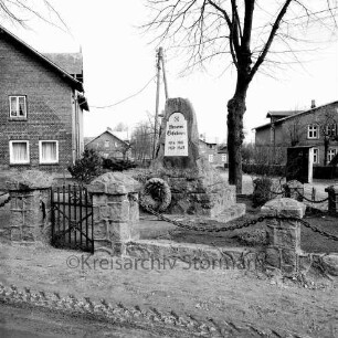 Hauptstraße: Ehrenmal mit Einfriedung und Baum: dahinter Bauernhäuser und Strommast