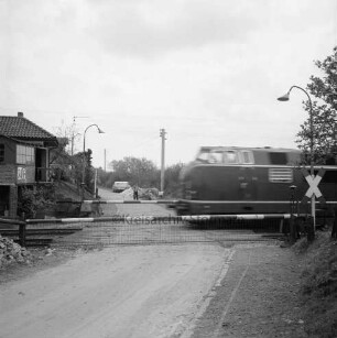 Bahnstrecke zwischen Hamburg und Lübeck: Bahnübergang an Schrankenposten 70 mit geschlossenen Schranken und Andreaskreuz: rechts durchfahrende Diesellok: im Hintergrund Arbeiter mit Schubkarre: dahinter Pkws