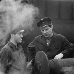 Zwei Eisenbahner zwischen Gleisen im Gespräch, umgeben von Dampfschwaden (einer Lokomotive), im Vordergrund ein Hülsenpuffer (Serie Deutsche Reichsbahn - Brigade "Otto Buchwitz")