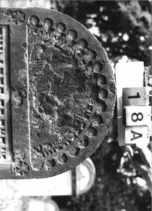 Grabstein 18 (Index: 100110924) Material: Sandstein Zustand: im Giebel Abschieferungen mit großem Textverlust, im unteren Teil leicht verwittert mit geringem Textverlust, eingesunken Maße HxBxT: 95x73x11 cm Beschreibung: Stele, Rundbogenabschluß mit Rundbogenfries, Schriffelder eingetieft Symbolik: 3 Rosetten: Grabstein 18A