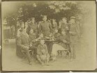 Garten mit stehender oder sitzender Tischrunde in Kriegsschule Engers: sieben Offiziere in Uniform teils mit Mütze, zwei Frauen, Kind mit Hund