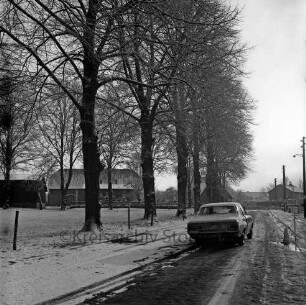 Kirchenweg mit Kopfsteinpflaster: rechts Pkw: links Grünfläche mit Bäumen: hinten Hauptstraße mit Häusern: Winter